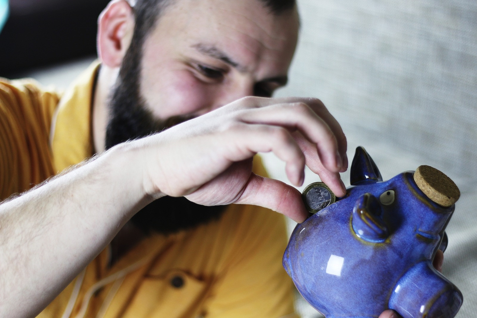 A bearded man in a yellow shirt placing a coin into a blue ceramic piggy bank while sitting indoors.