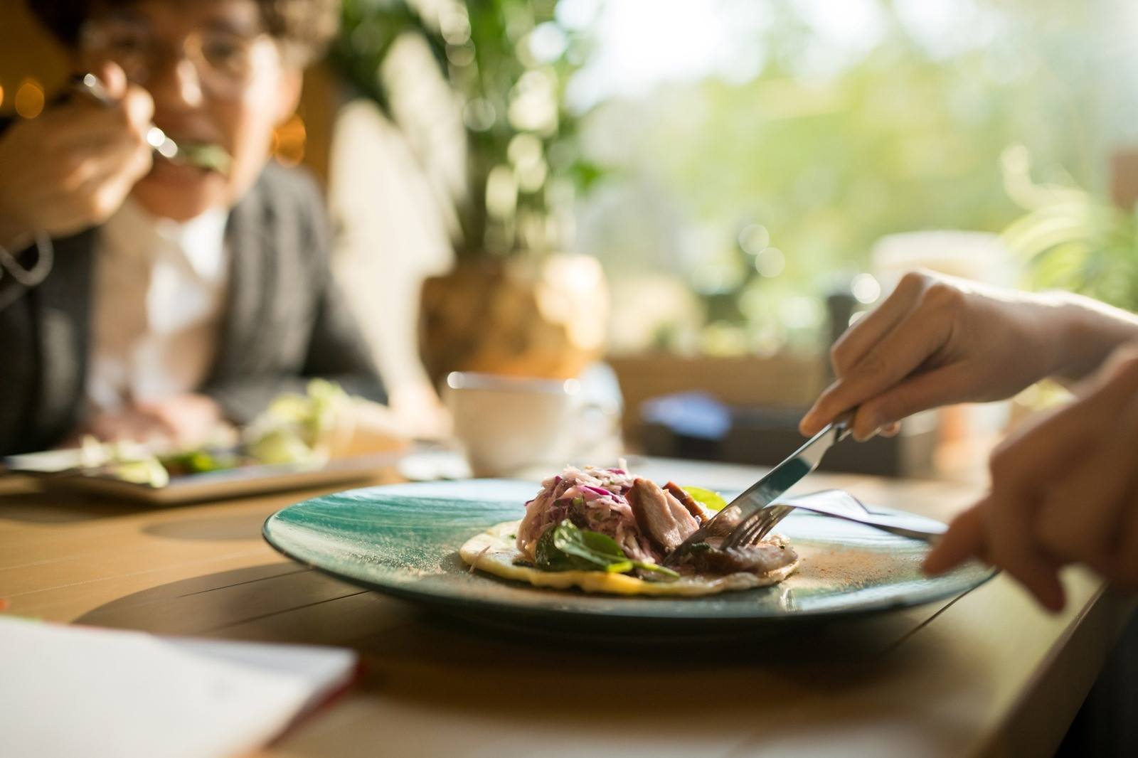 Close-up of a person cutting food on a plate at a table while another person eats in the background.