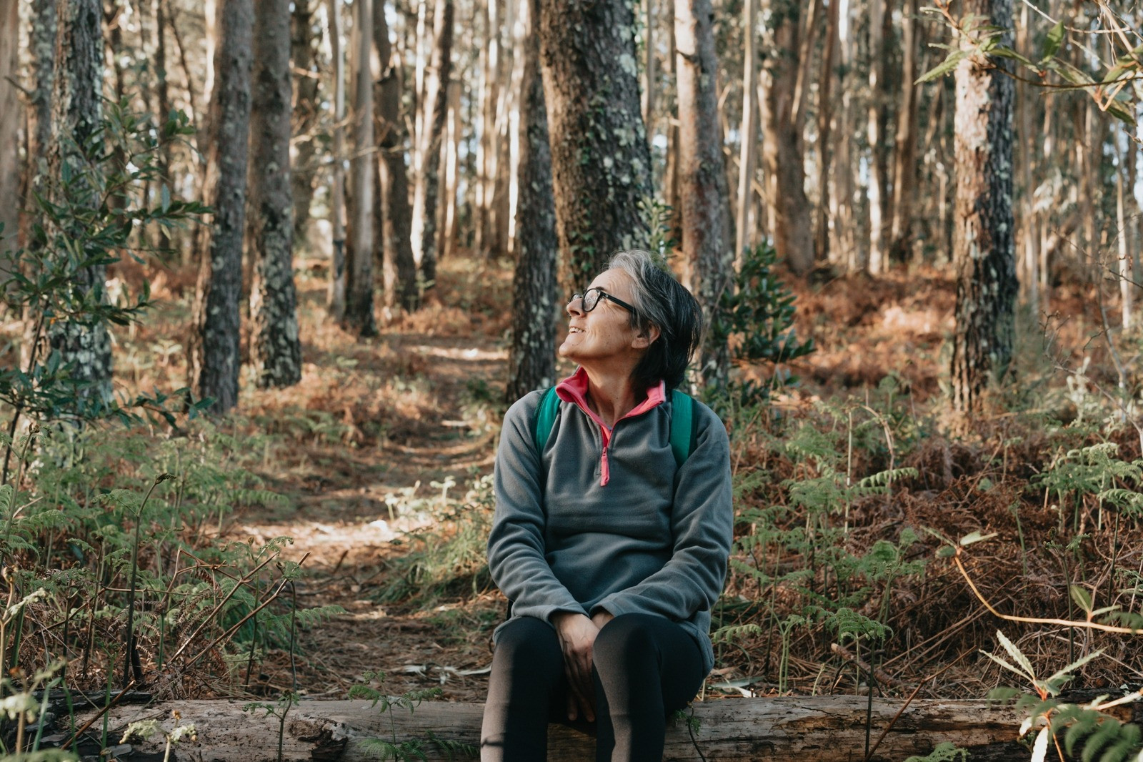 Woman with a backpack sitting on a fallen log in a wooded area and looking upward.