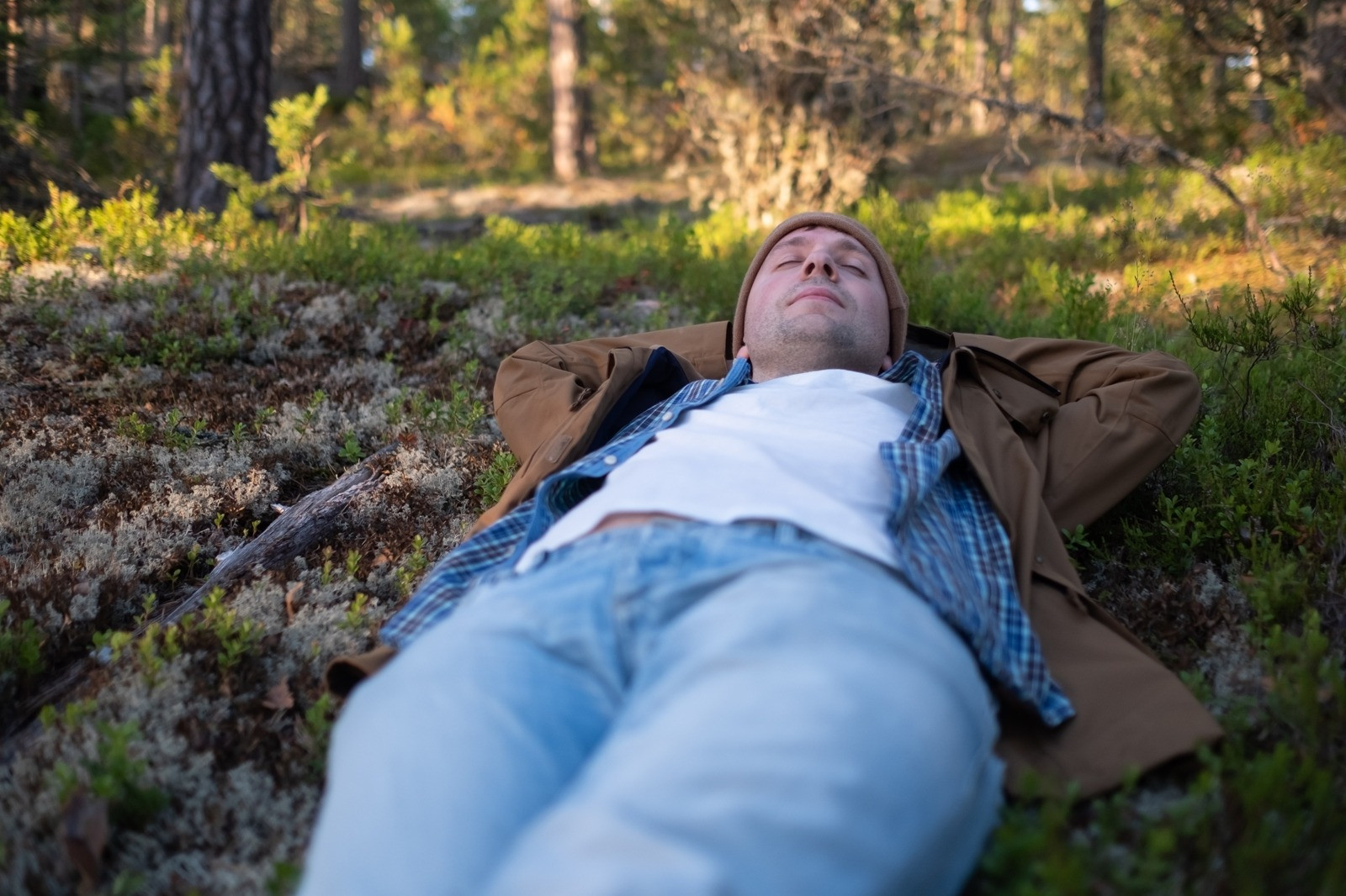 Man lying on the forest floor with his eyes closed and hands behind his head in a calm outdoor setting.