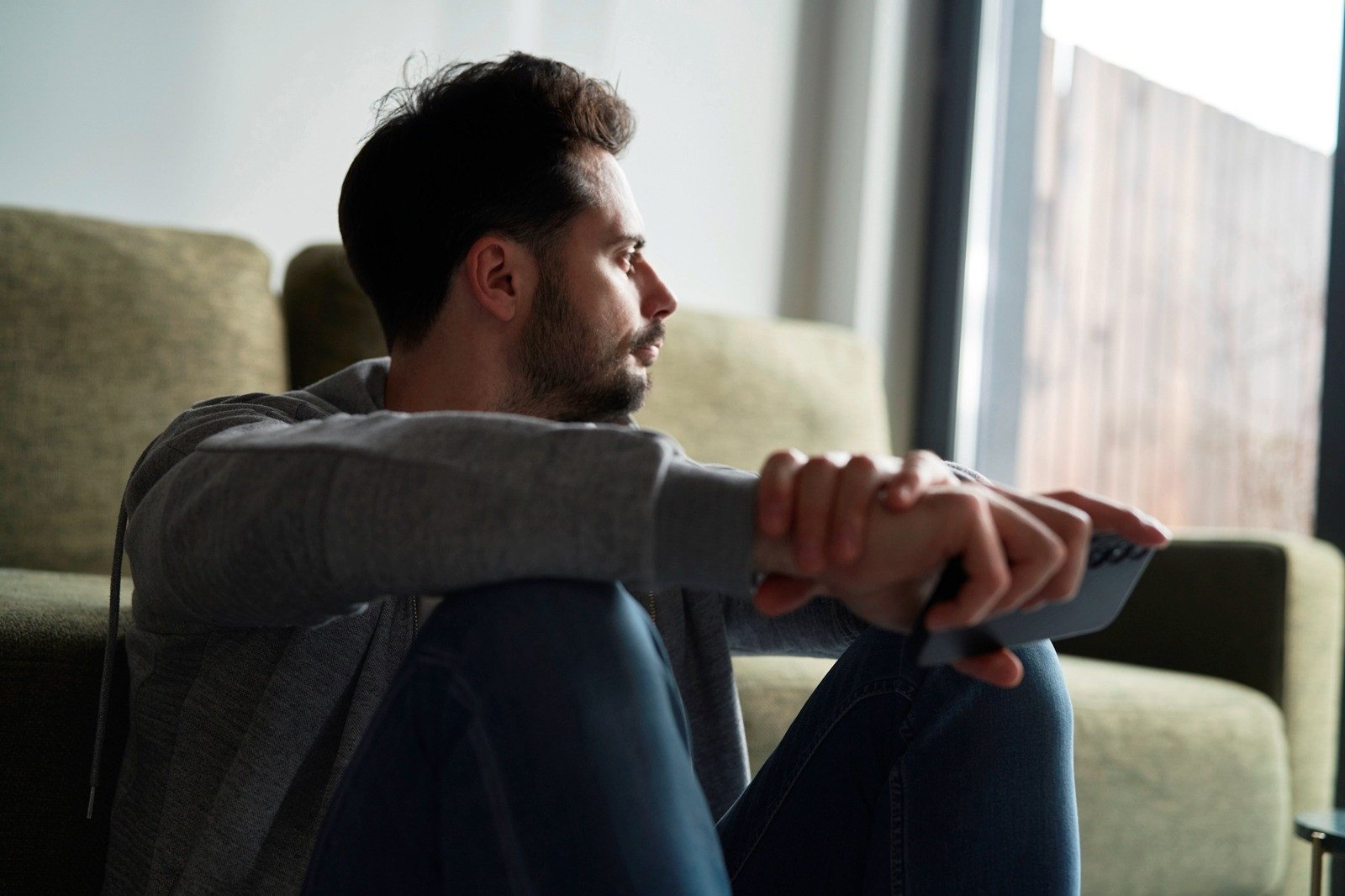 Man sitting on a sofa near a window, resting his arms on his knees and looking thoughtfully outside.