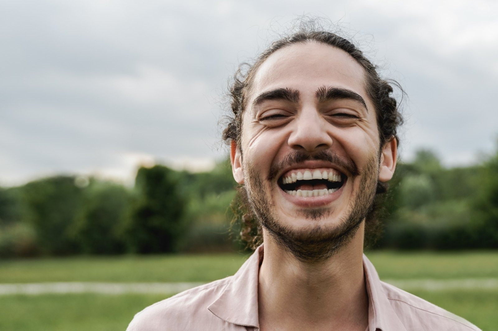 Smiling man with dark curly hair and a beard wearing a light pink shirt outdoors against a green landscape.