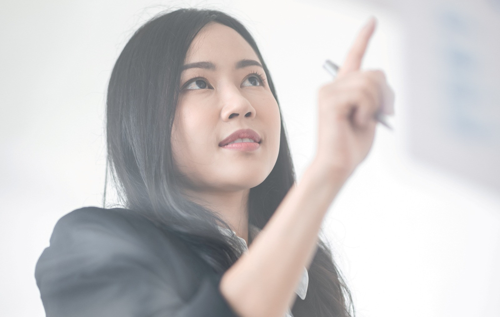 Woman looking upward and pointing with a pen toward a glass surface in a bright, softly lit setting.
