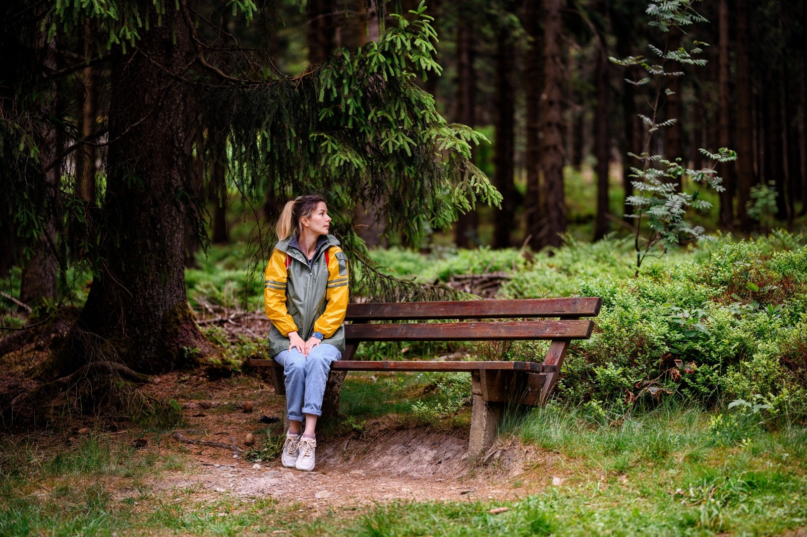 Woman sitting alone on a bench in a forest, looking to the side in a quiet, reflective moment.