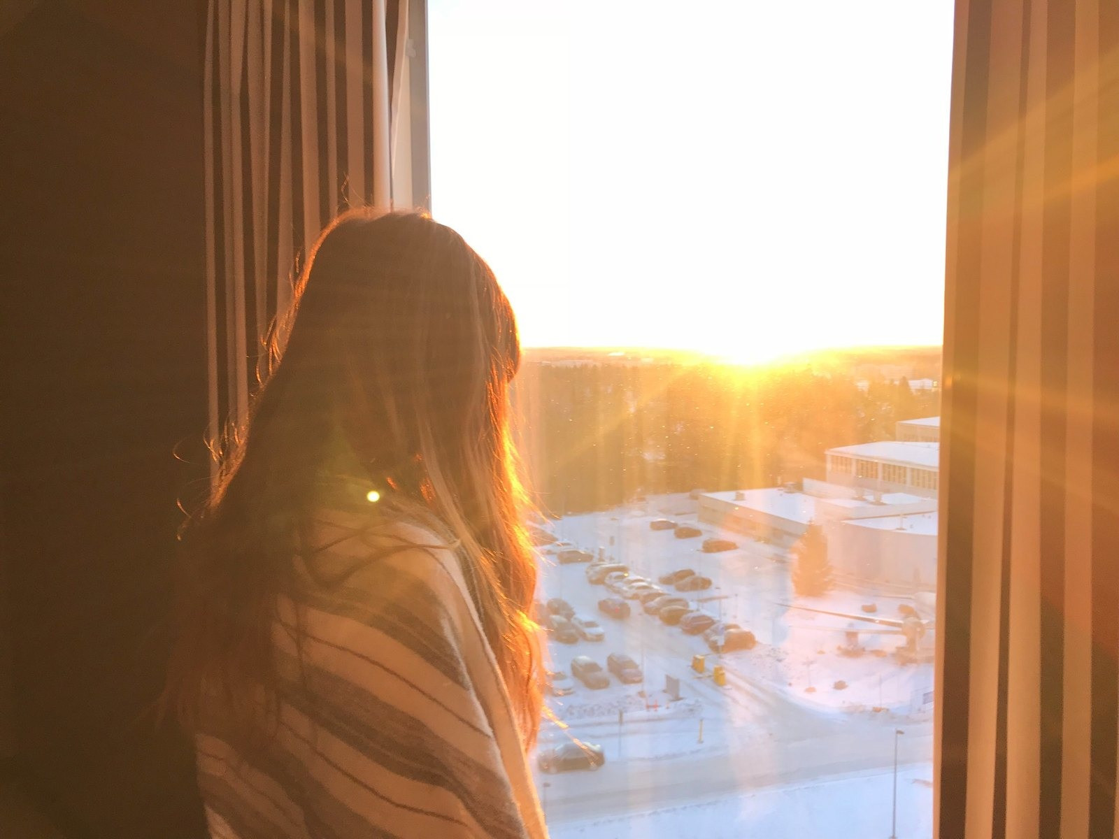 Woman looking through a window toward a sunset over a snow-covered landscape and buildings.