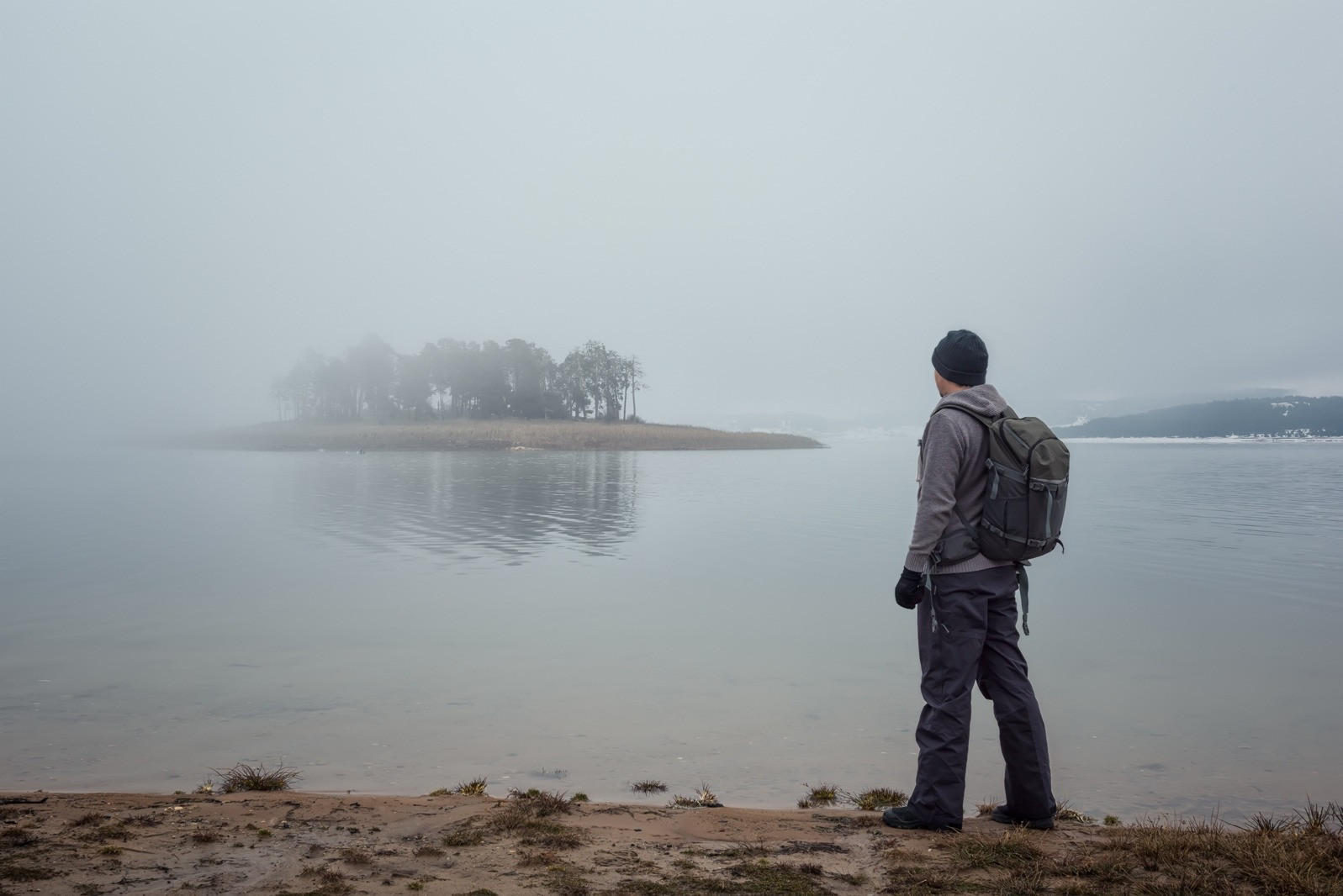 Man wearing a backpack standing beside a calm, misty lake, looking toward a distant island surrounded by fog.
