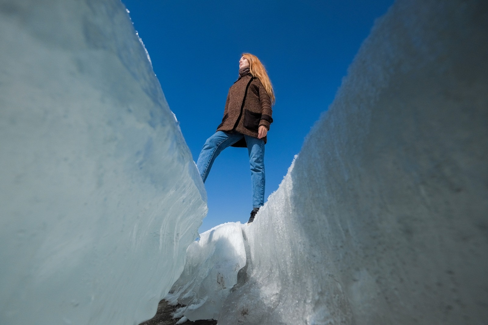 Woman standing between tall walls of snow beneath a clear blue sky, viewed from below.